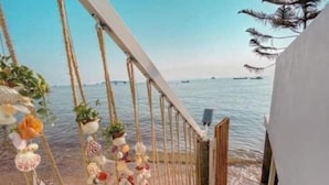 Eine Holzterrasse mit Geländer, verziert mit Meeresschalen und kleinen Pflanzen, mit Blick auf ein ruhiges Meer und in der Ferne Boote.