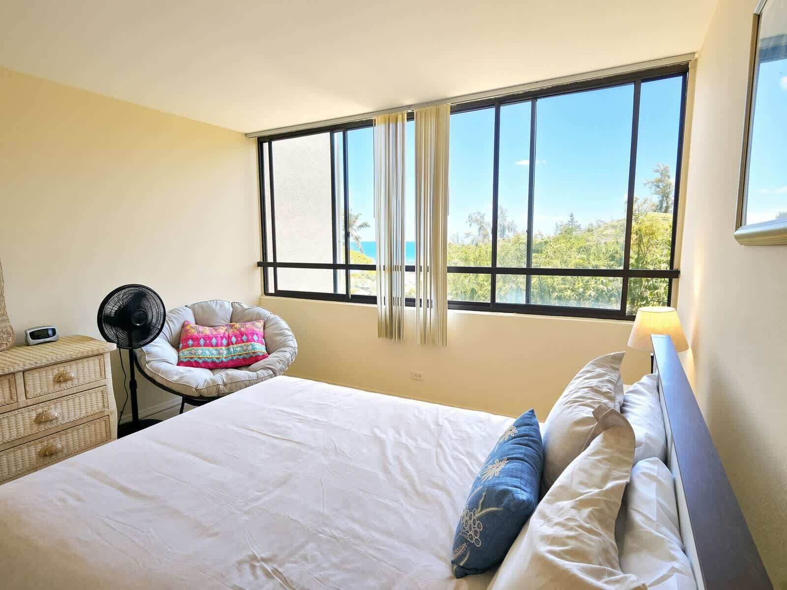 A bedroom with a bed, a chair, a fan, a dresser, and a window with a view.