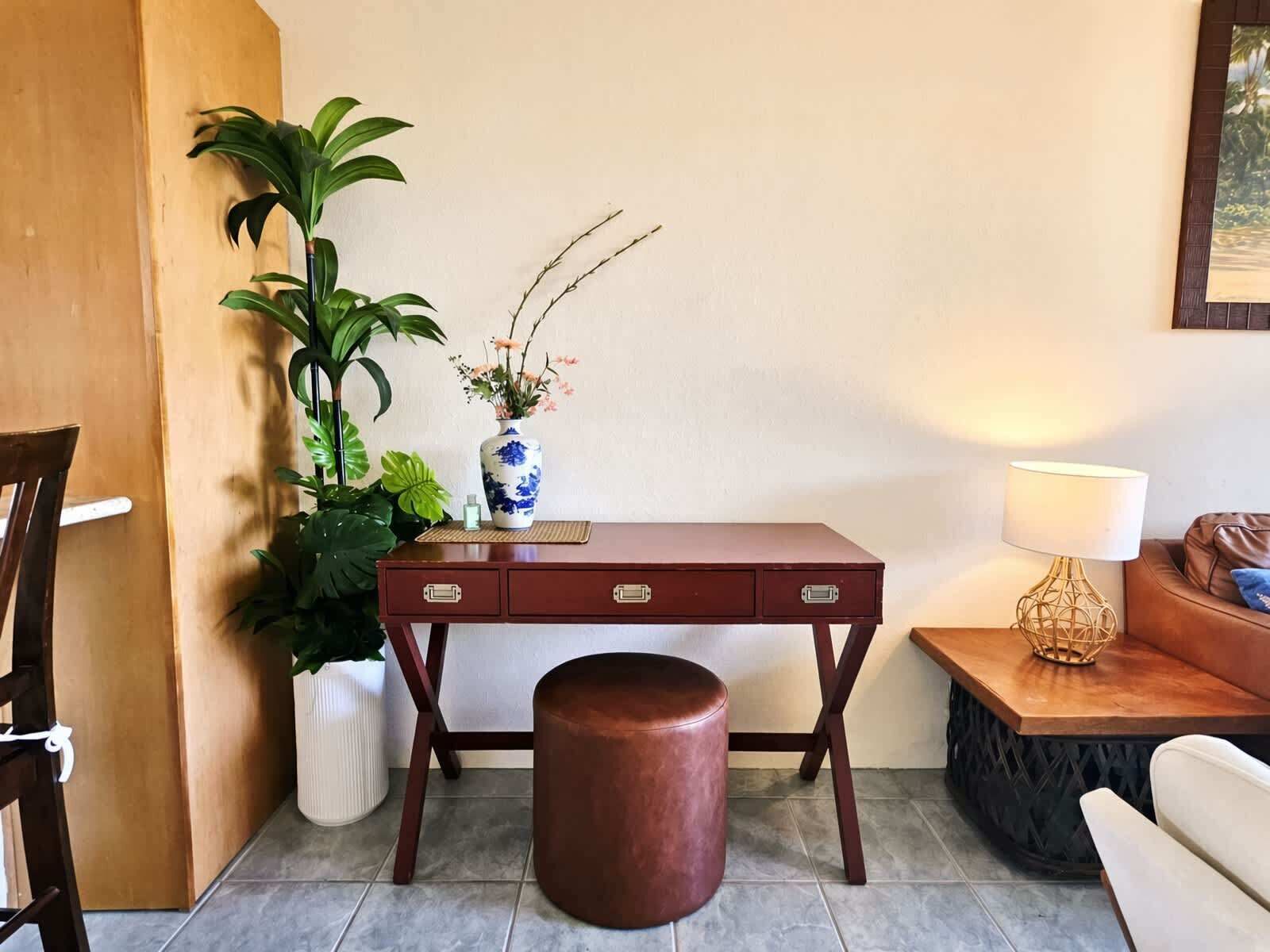 Quarto com escrivaninha de madeira, banquinho acolchoado, luminária e plantas em vasos.