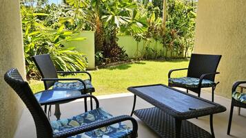 An outdoor patio area with wicker furniture and a glass table, surrounded by lush tropical plants.