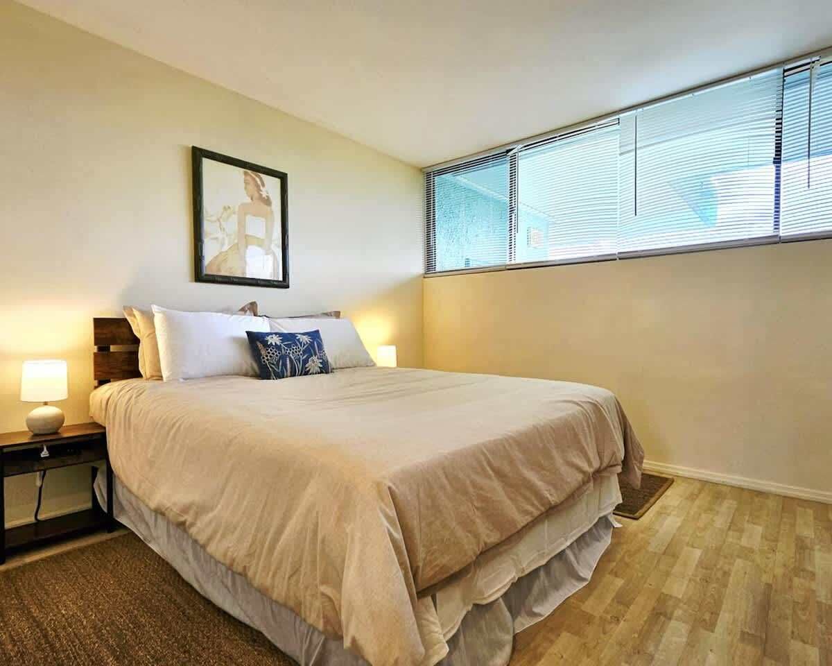 A bedroom with a bed, a nightstand, a lamp, a window with blinds, and a framed picture on the wall.