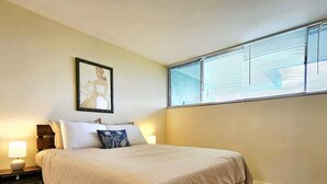A bedroom with a bed, a nightstand, a lamp, a window with blinds, and a framed picture on the wall.
