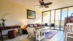 A living room with a sofa, coffee table, and a desk. There is a ceiling fan, a painting on the wall, and a view through a glass door.