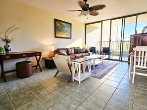 A living room with a sofa, coffee table, and a desk. There is a ceiling fan, a painting on the wall, and a view through a glass door.