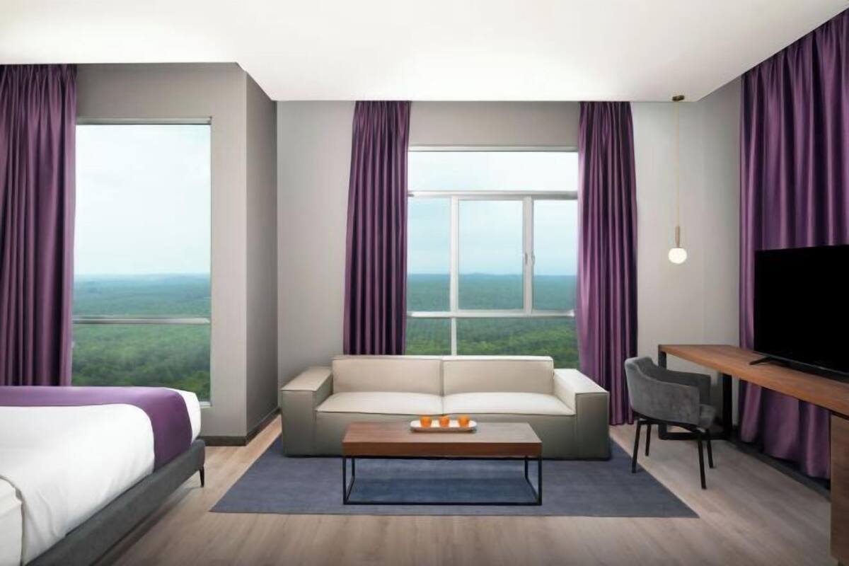 A modern hotel room with a large window, a sofa, a coffee table, a TV, and a desk.