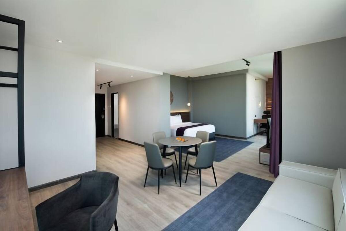 A modern hotel room with a bed, a dining table with chairs, a sofa, and a small kitchen area.