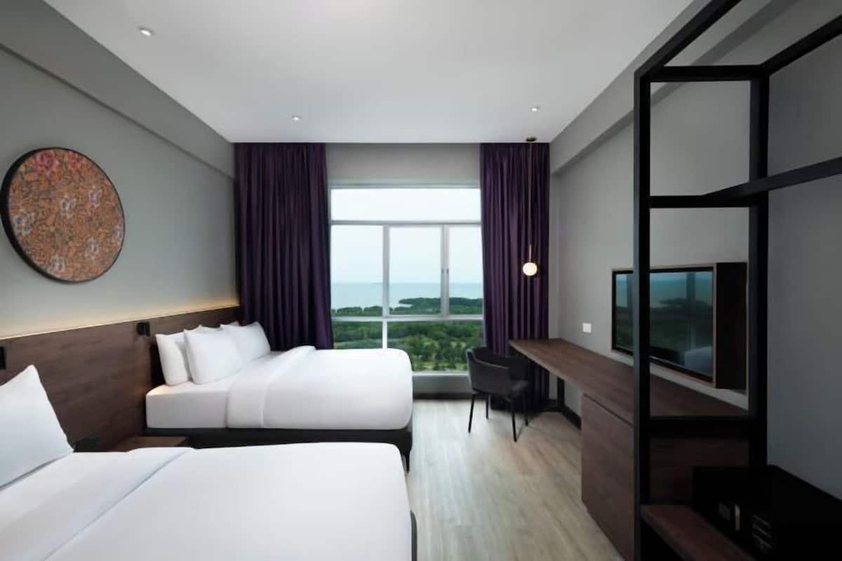A hotel room with two beds, a desk, a TV, and a large window offering a view of the outdoors.