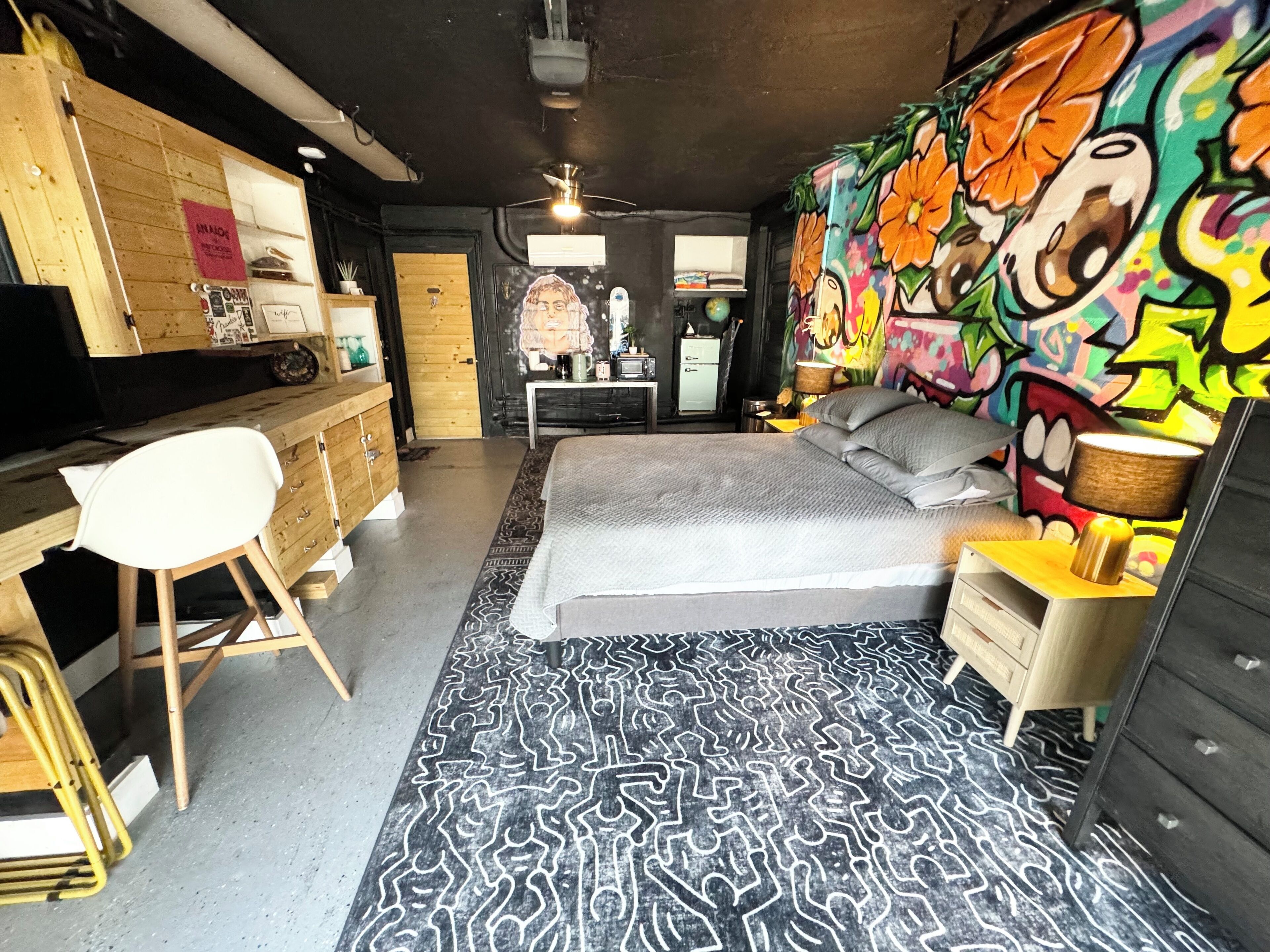 Une chambre à coucher comprenant un lit, un bureau, une chaise, une commode et une fresque colorée sur le mur.