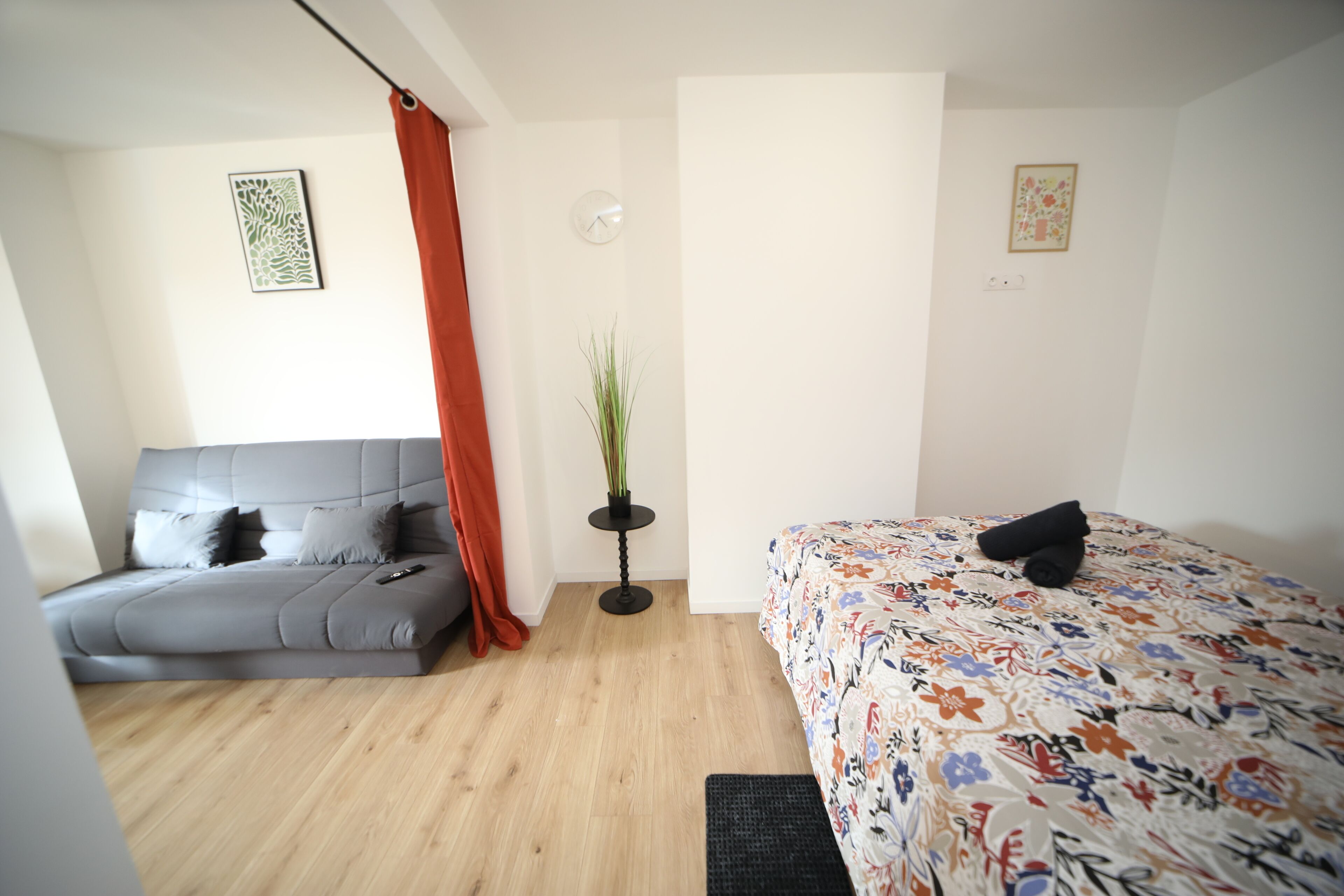 Een moderne slaapkamer met een grijze bank, een bloemenbedekking, een potplant en een rood gordijn.