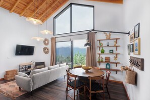 Uma sala de estar moderna com uma janela grande, uma TV de tela plana, um sofá, uma mesa de jantar com cadeiras e prateleiras decorativas.