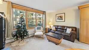 Ein gemütliches Wohnzimmer mit Weihnachtsbaum, einem braunen Ledersofa, einem Holztisch und einem großen Fenster mit Blick auf Gebäude.