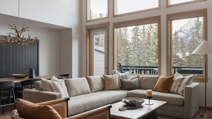 Amplia sala de estar con un sofá seccional grande, una mesa de centro y un comedor con mesa y sillas de madera. Amplias ventanas ofrecen vistas al exterior.