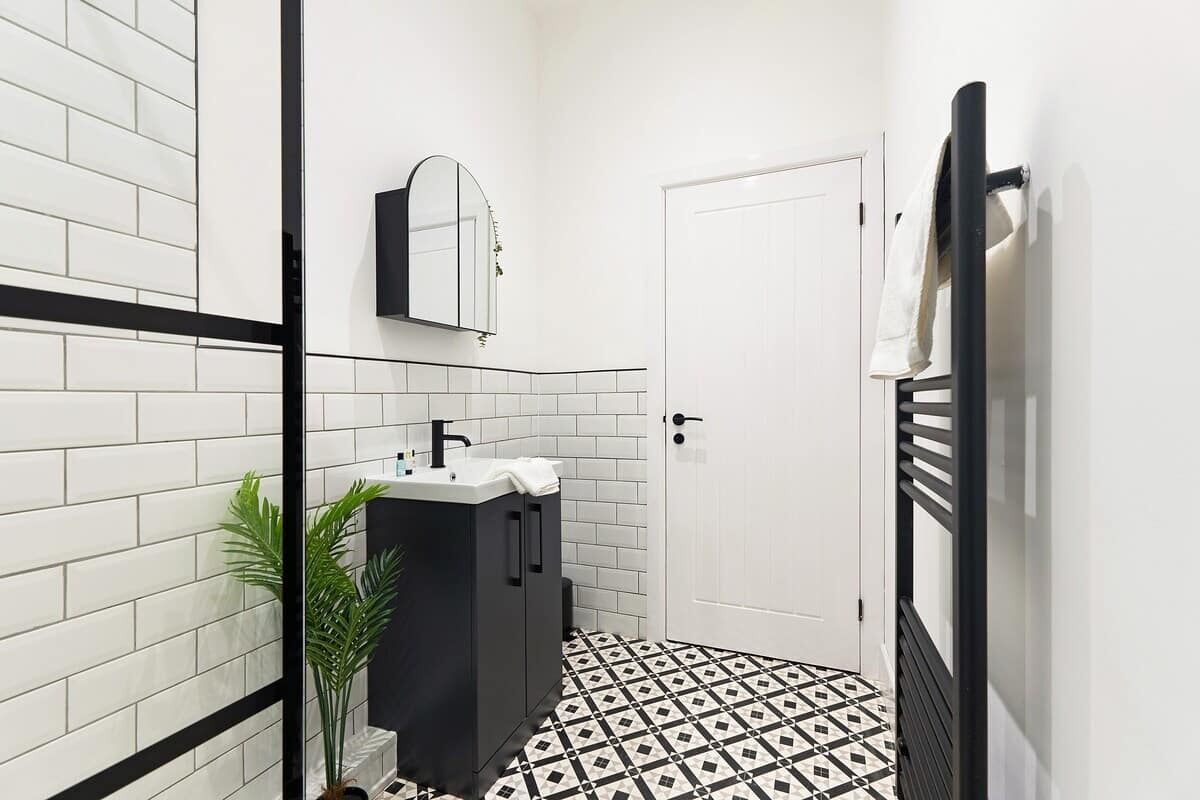 A bathroom with white tiled walls, a black and white patterned floor, a black cabinet with a sink, a mirror, and a towel rack.