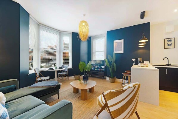 Una sala de estar moderna con un sofá, una silla a rayas, una mesa de centro redonda y una zona de cocina con encimera blanca y una lámpara colgante.