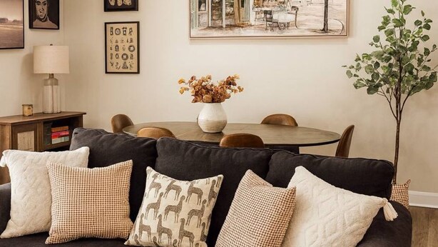 A living room with a dark sofa, a coffee table, and a dining table with chairs. There is a painting on the wall, a vase with dried flowers, and a potted plant. - Charming 3-bedroom house with AC, fitness room in fabulous Greenville (Greenville)