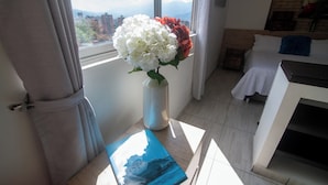 A room with a bed, a window with curtains, a vase with flowers, and a book on a table. - Staynova Comfortable Loft in El Poblado (Medellín)