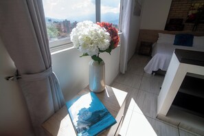 A room with a bed, a window with curtains, a vase with flowers, and a book on a table. - Staynova Comfortable Loft in El Poblado (Medellín)