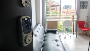 A room with a Yale smart lock on a door, a grey sofa, a red chair, and a view of buildings through a window. - Staynova Comfortable Loft in El Poblado (Medellín)