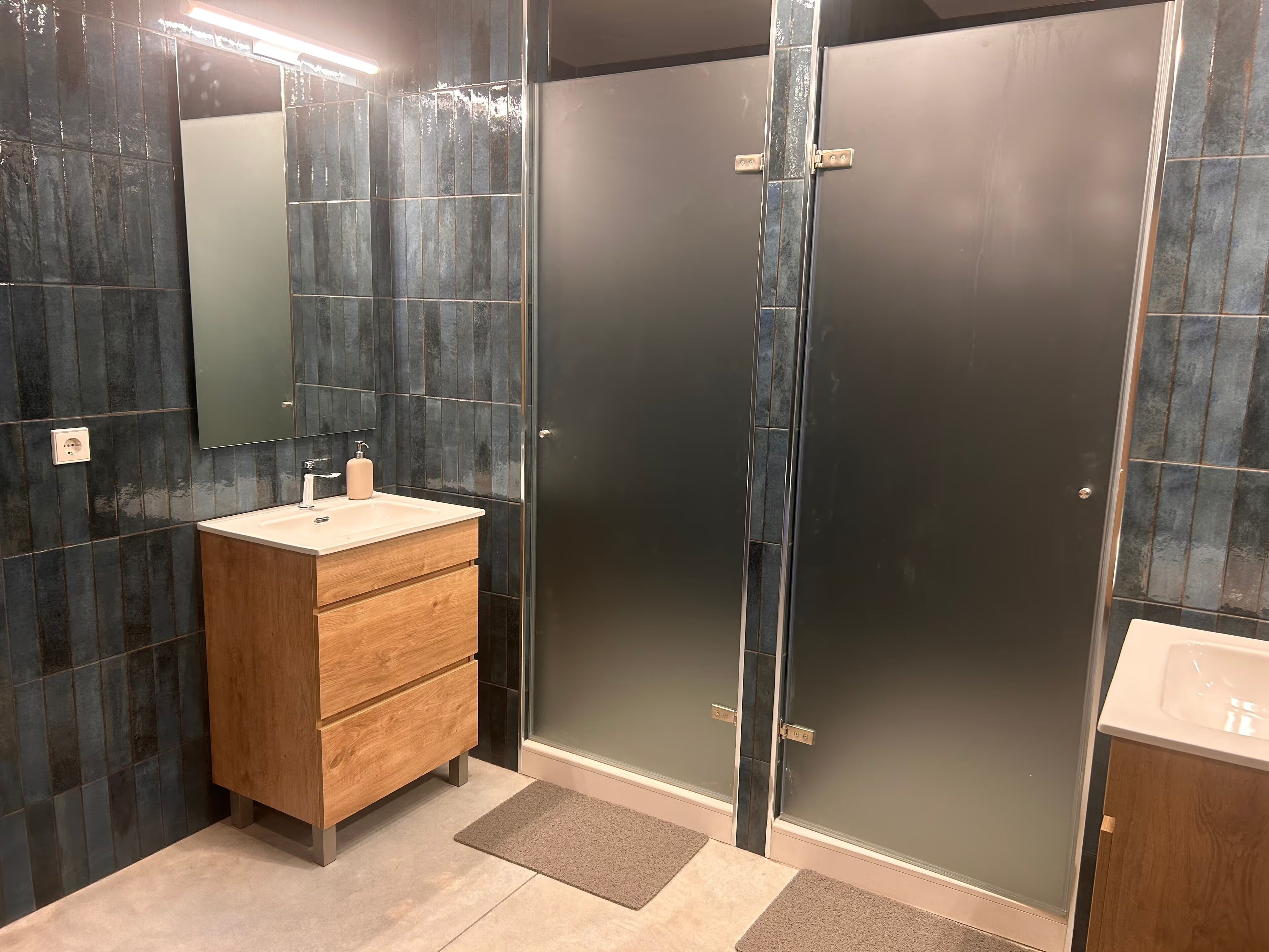 Un baño moderno con paredes de azulejos oscuros, un tocador de madera con un lavamanos y una cabina de ducha con vidrio esmerilado.