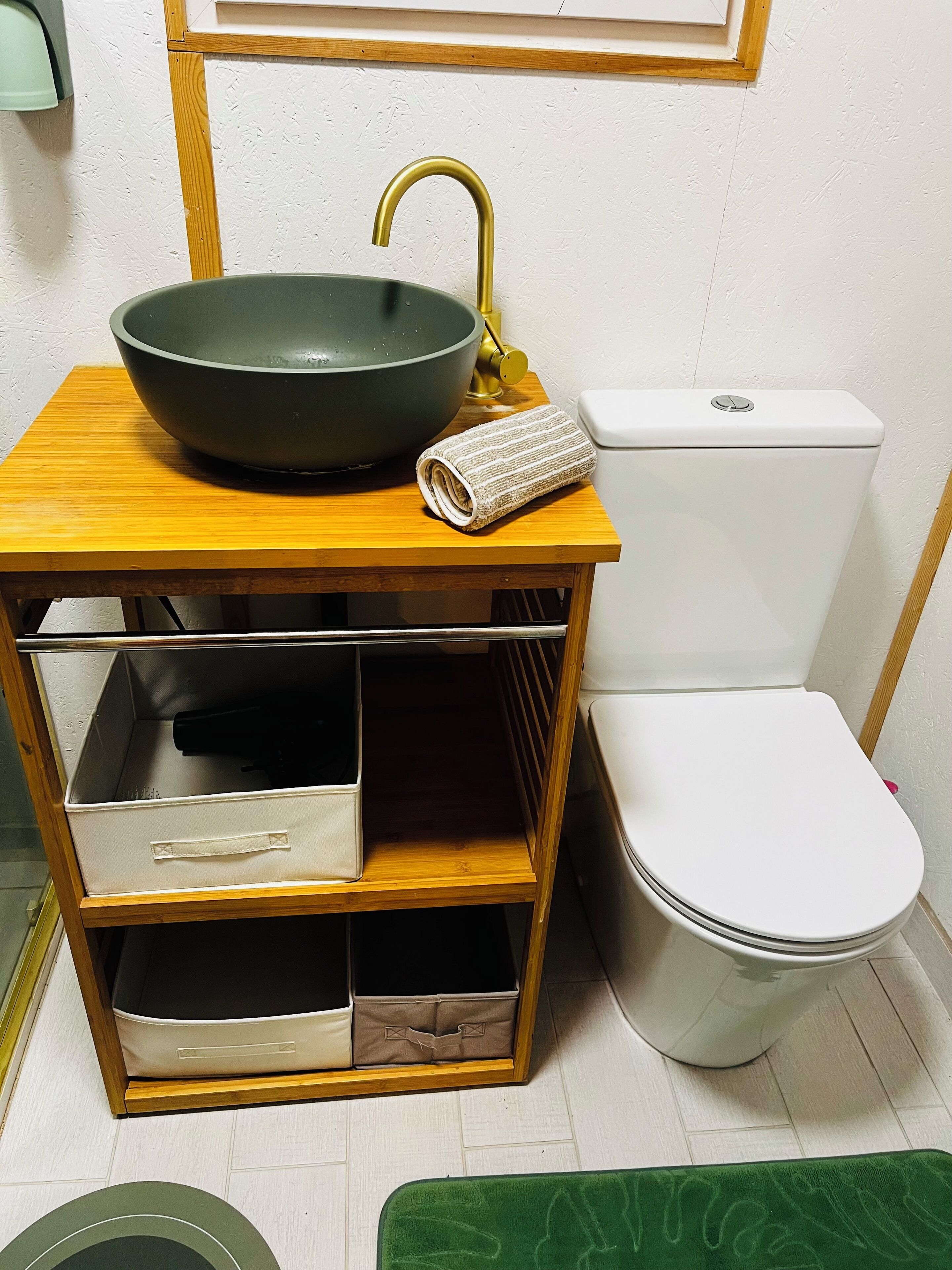 A compact bathroom with a wooden vanity, a large bowl sink, a toilet, and a green mat.