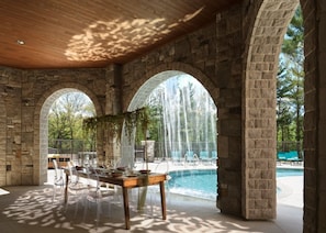 A stone archway leading to a pool with a waterfall, clear chairs, and a wooden table with a view. - Dellshire Resort (Wisconsin Dells)