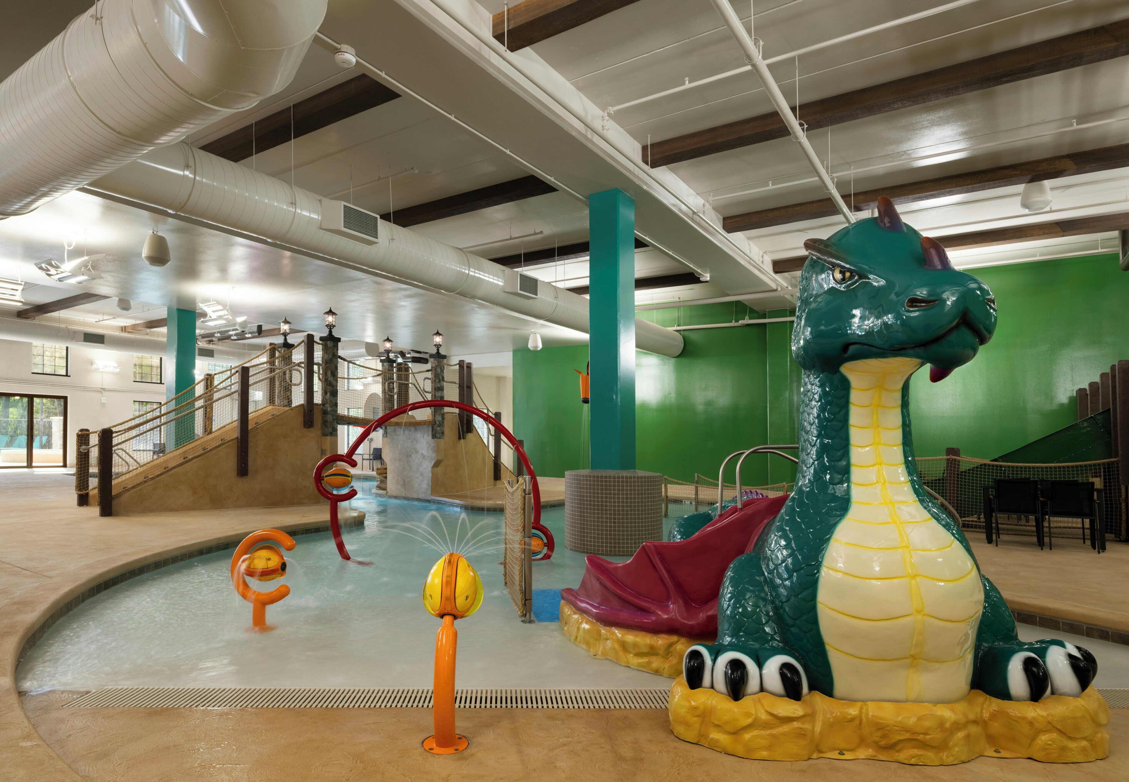 An indoor swimming pool with a large dinosaur sculpture, slides, and a netted climbing area.