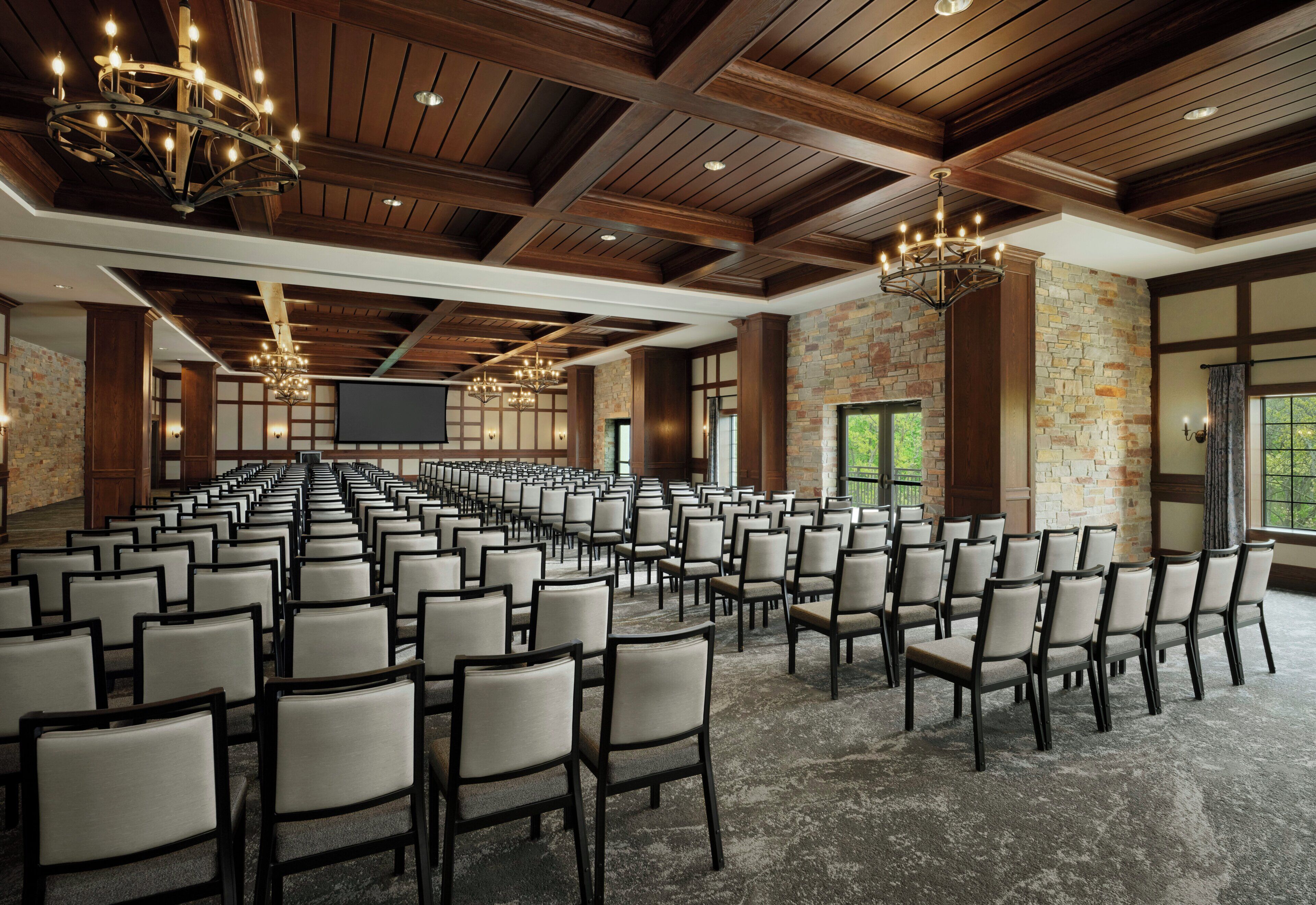 Un salón de conferencias con filas de sillas, vigas de techo de madera, paredes de piedra y candelabros.