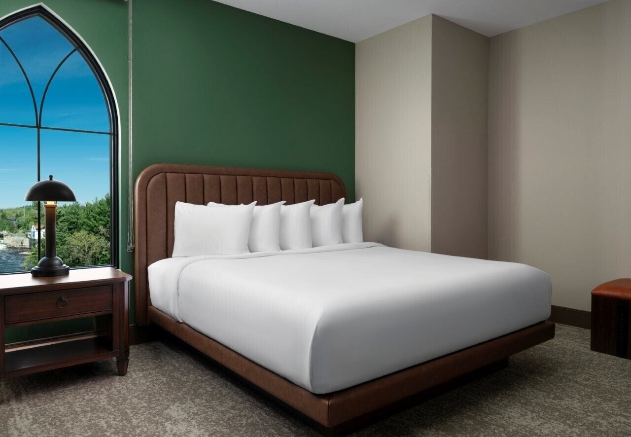 A bedroom with a large window, a bed with white linens, a wooden nightstand, and a lamp.