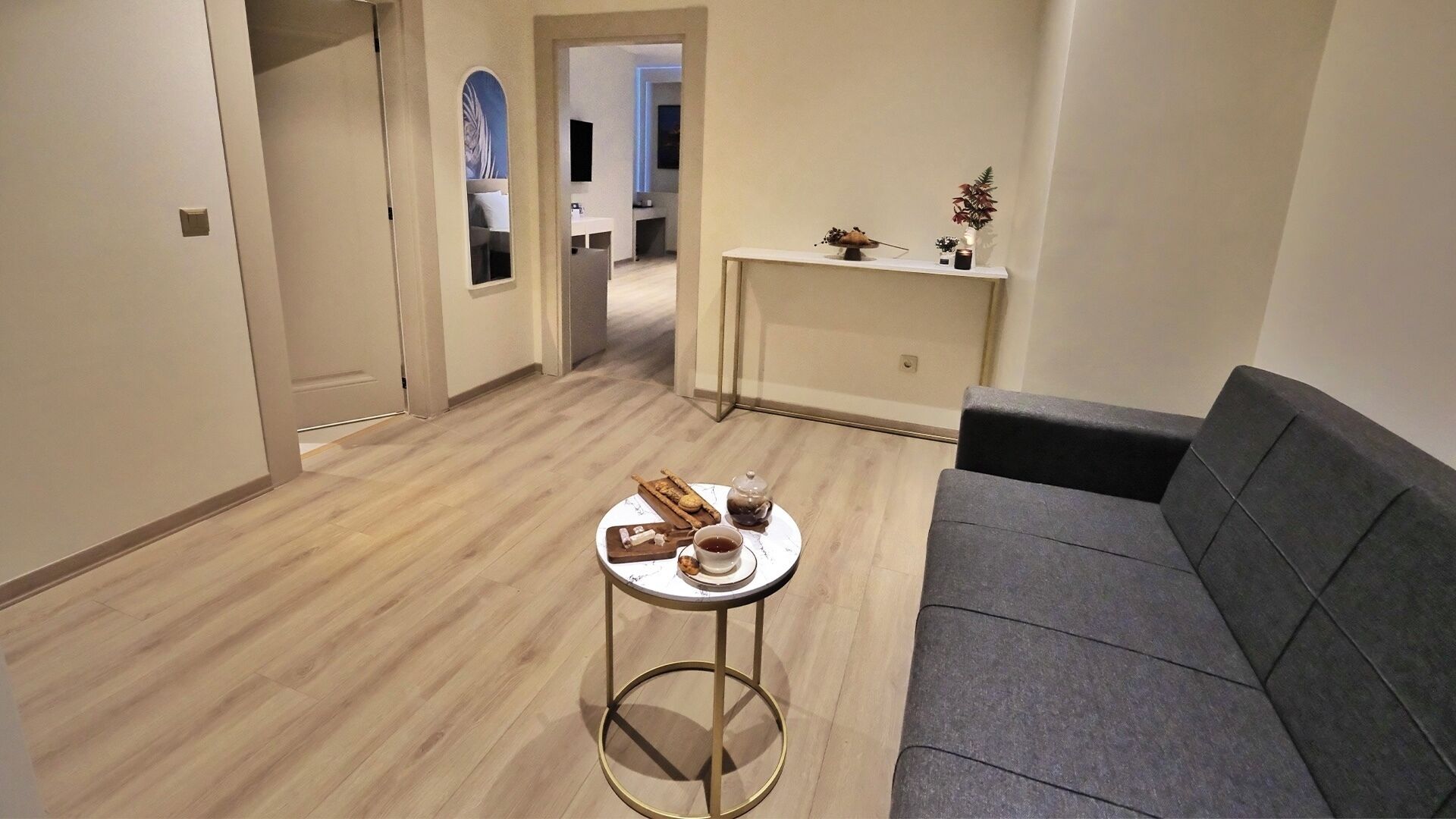 A modern living room with a grey sofa, a round coffee table with snacks and a teapot, and a hallway leading to another room.