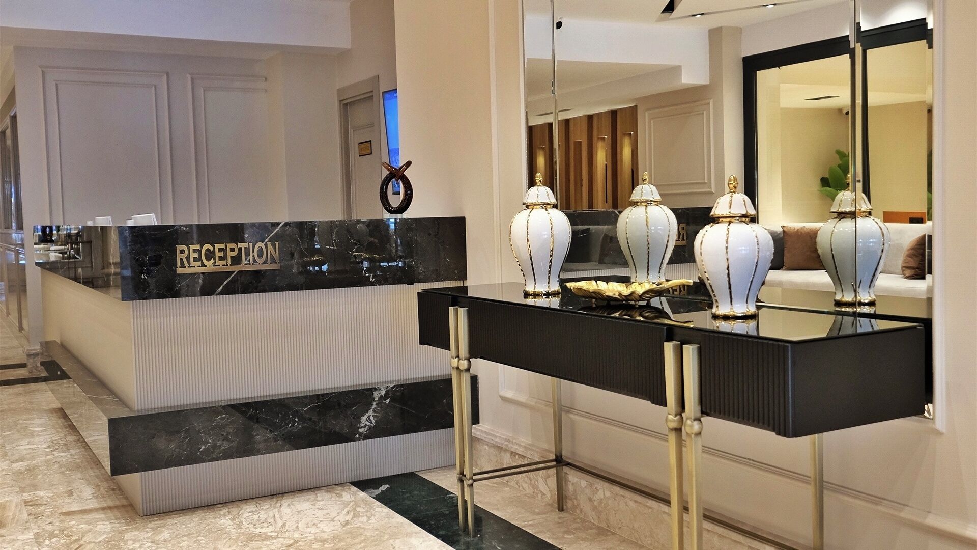 Un mostrador de recepción con encimera de mármol y detalles dorados, un espejo que refleja la sala, y un área de recepción con un sofá y jarrones decorativos.
