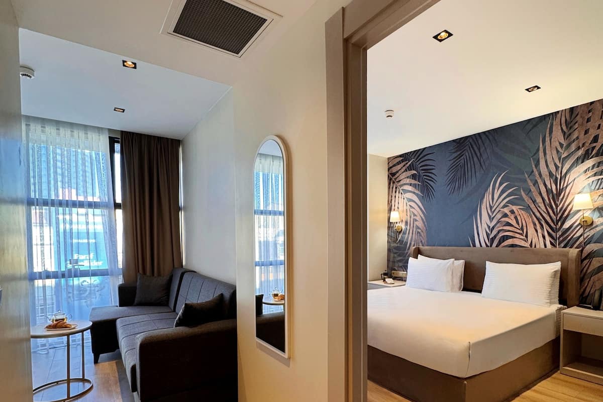 A modern hotel room with a large bed, a sofa, a small table, and a view of the city through large windows.