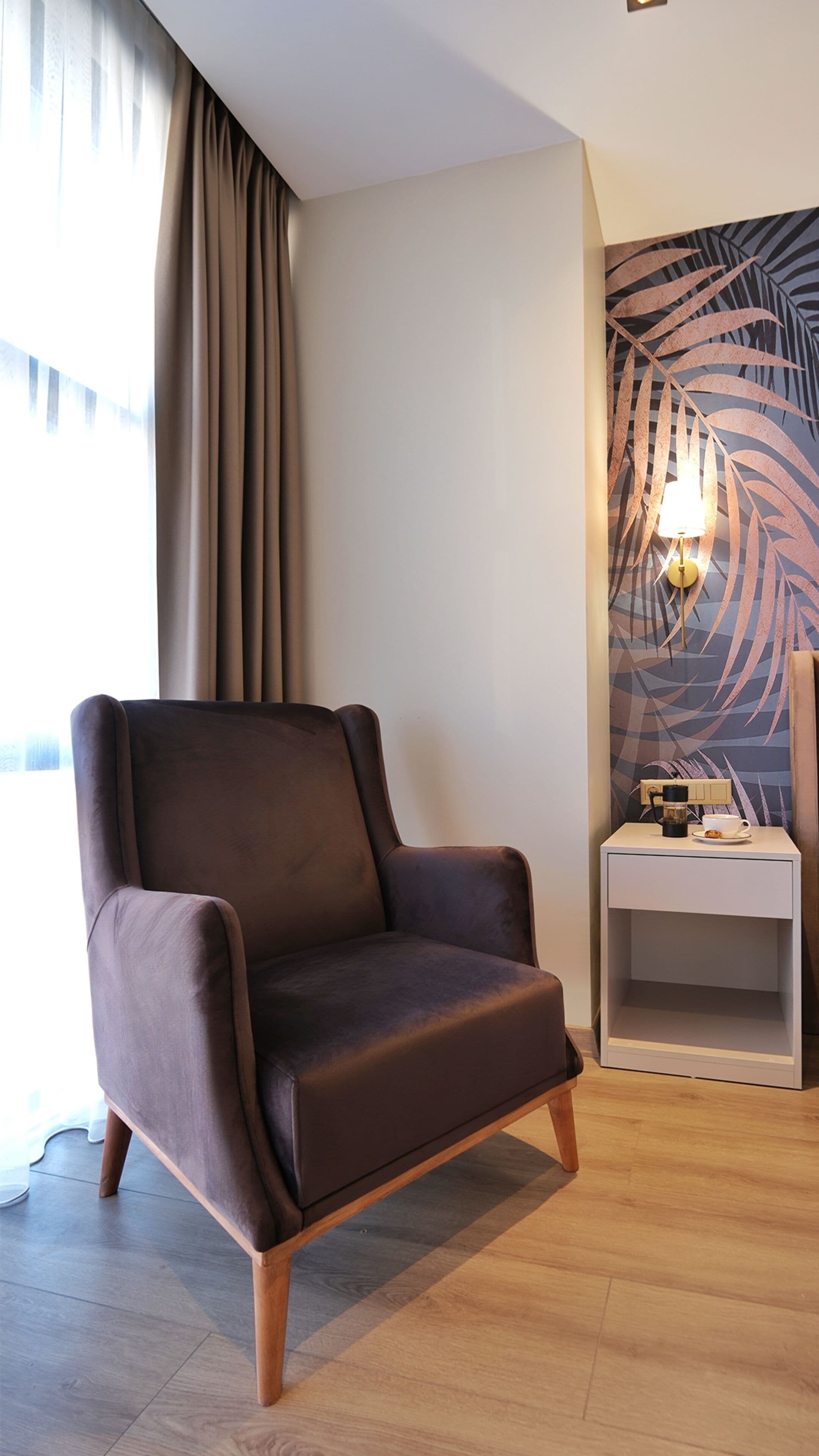 A modern room with a dark brown leather armchair, a small white nightstand with a lamp, and a wall with a leaf pattern.