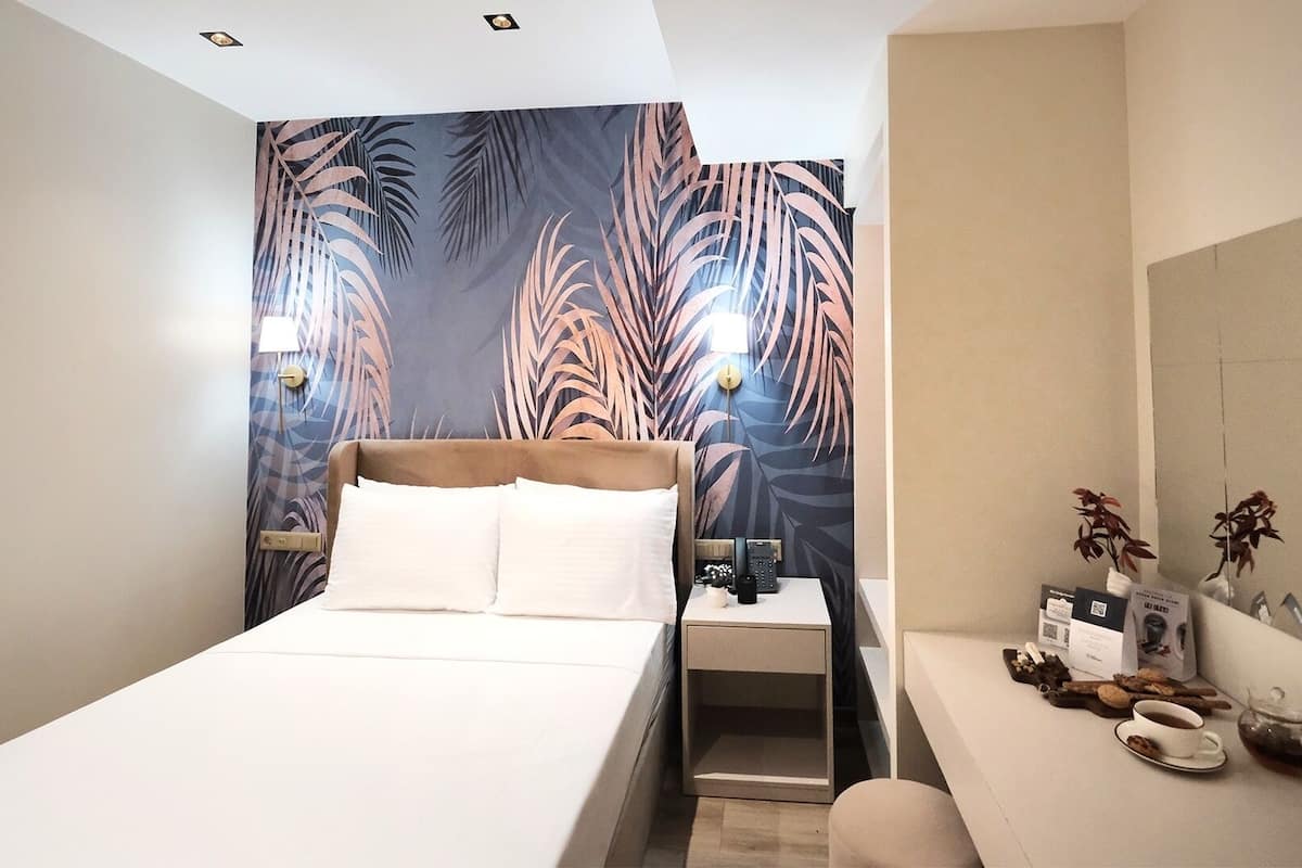 A modern hotel room with a large bed, a bedside table with a lamp, a small table with a vase and a cup, and a mirror on the wall.