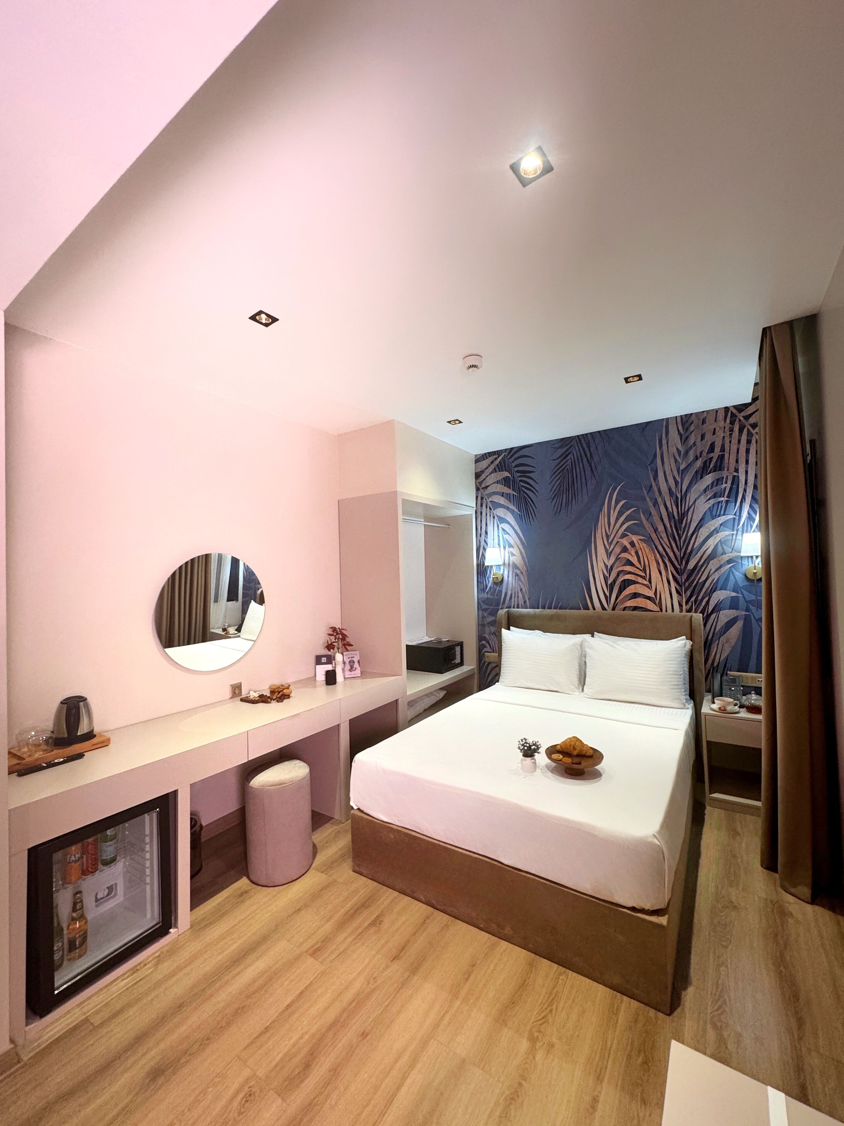 A modern hotel room with a large bed, a bedside table, a mirror, a small seating area, and a wall with a leaf pattern.