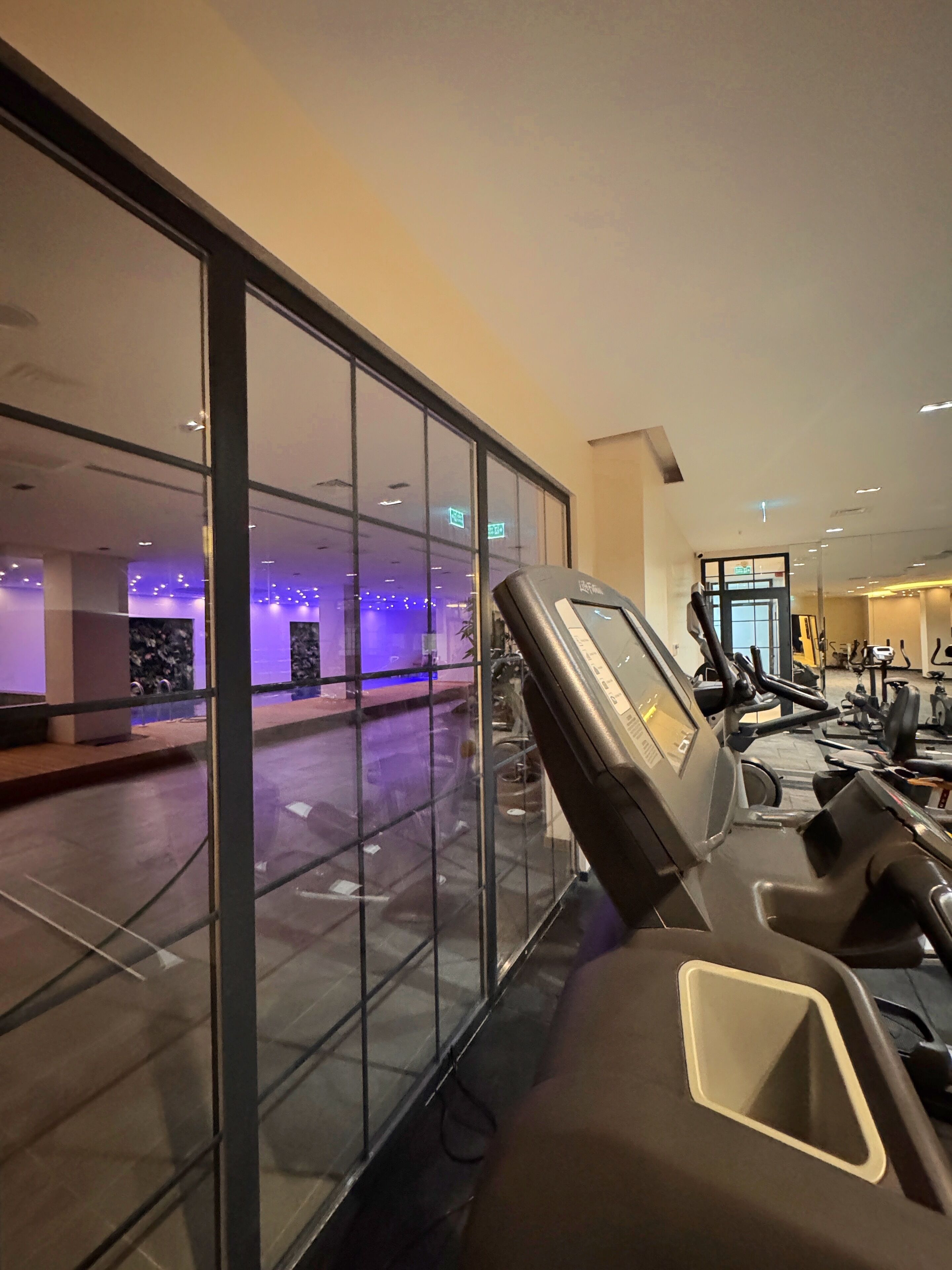 Une salle de sport équipée de matériel d’exercice, avec un grand mur en verre et un espace éclairé de lumière violette à l’arrière-plan.