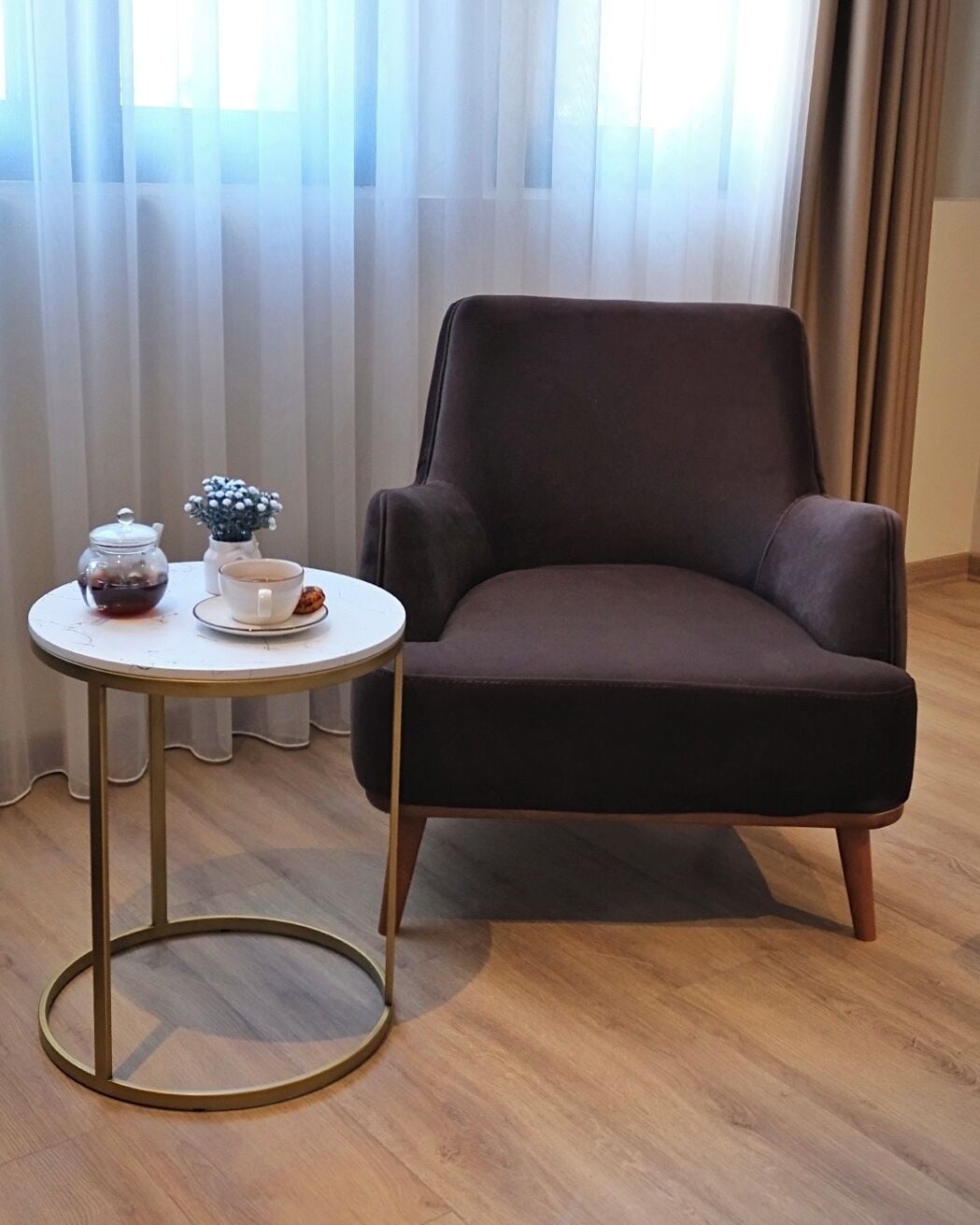 A dark brown armchair with a round table beside it, featuring a glass teapot, a white cup, and a small blue and white decorative item.