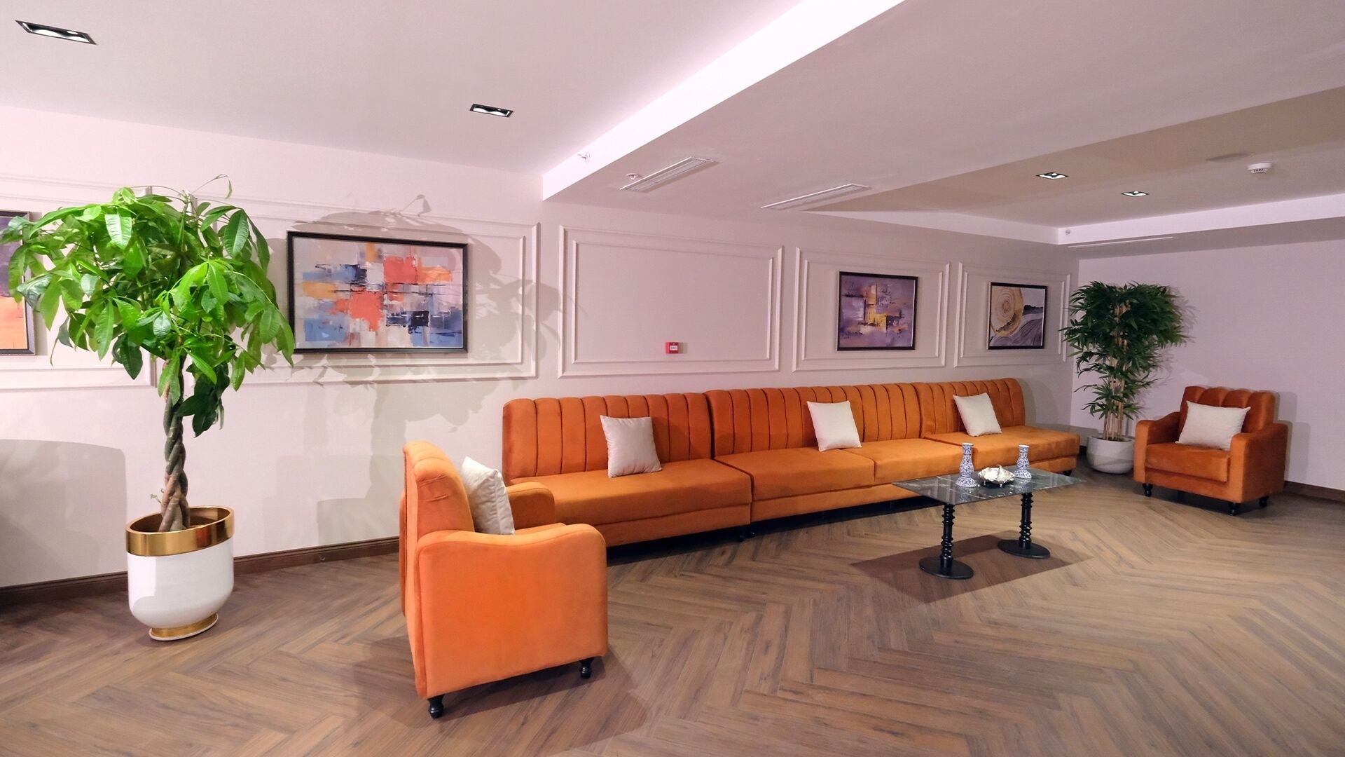 A modern lobby with a herringbone wood floor, an orange sofa set, a white armchair, and a large potted plant.