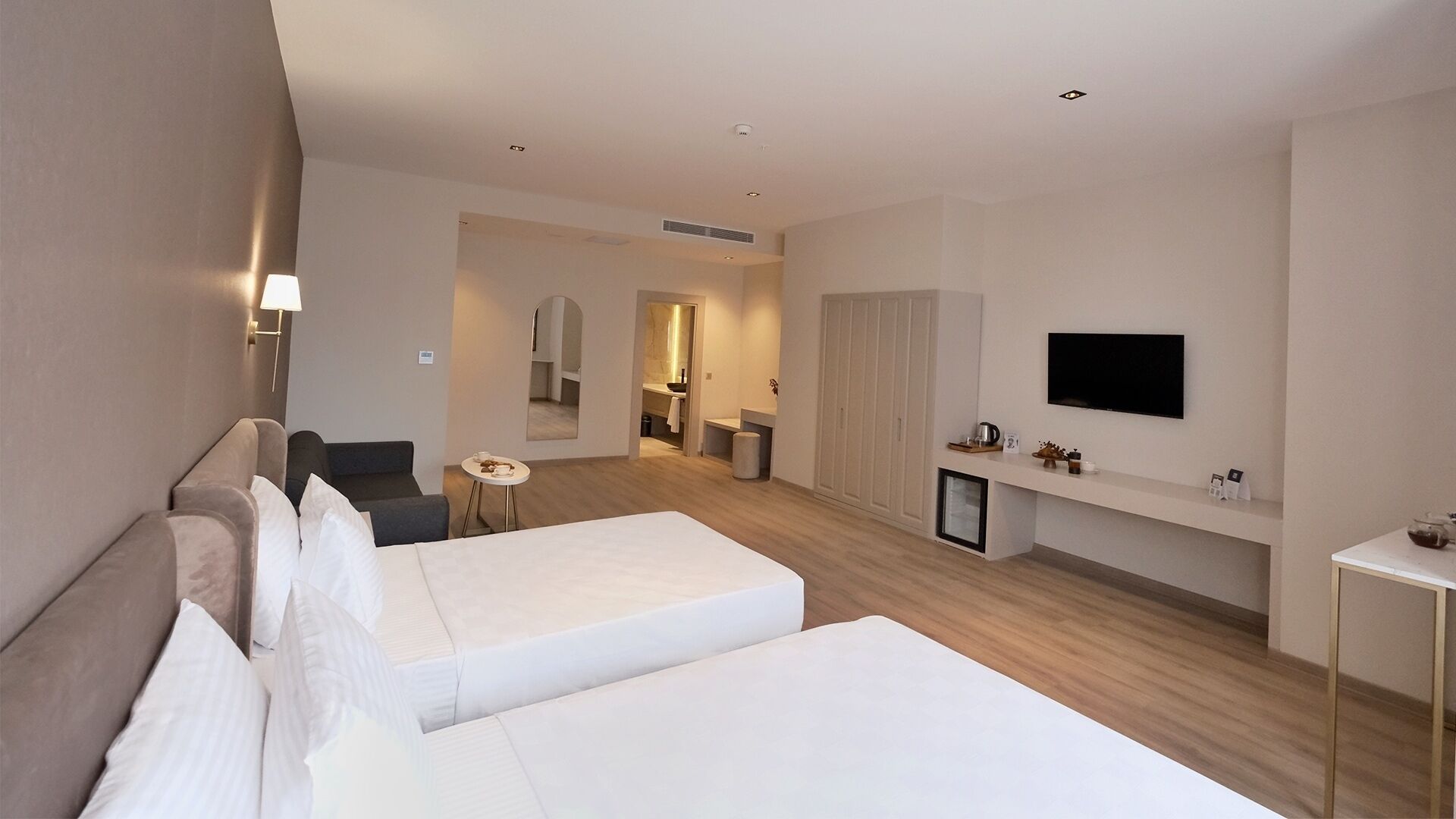 A modern hotel room with two beds, a TV, a small table, and a bathroom visible through an archway.