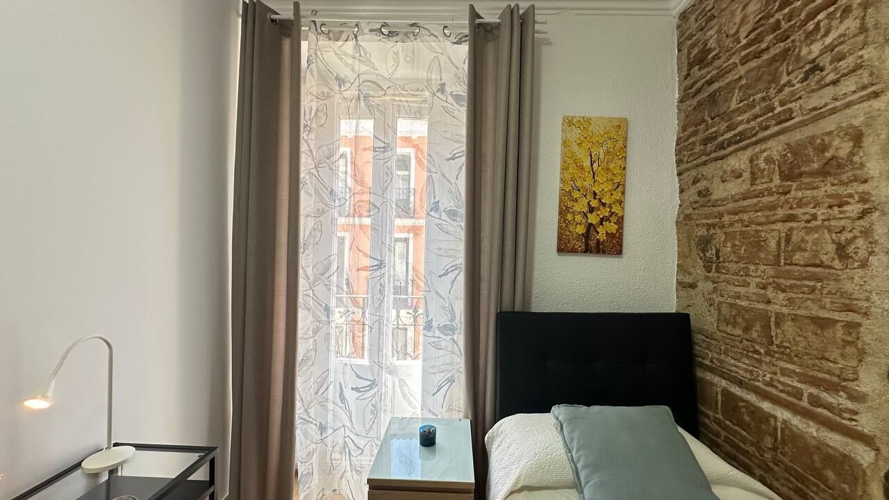 Un dormitorio con cama, mesita de noche, lámpara, ventana con cortinas y pared de ladrillo.