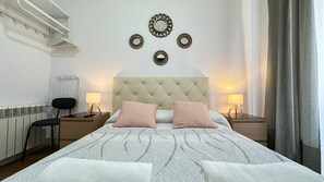A neatly arranged bedroom with a tufted headboard, two bedside lamps, a radiator, a chair, and decorative wall mirrors. - Habitaciones Manuela (Madrid)