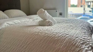 Una cama bien hecha con una toalla doblada en el centro, una ventana con vistas a edificios y una pared sencilla.
