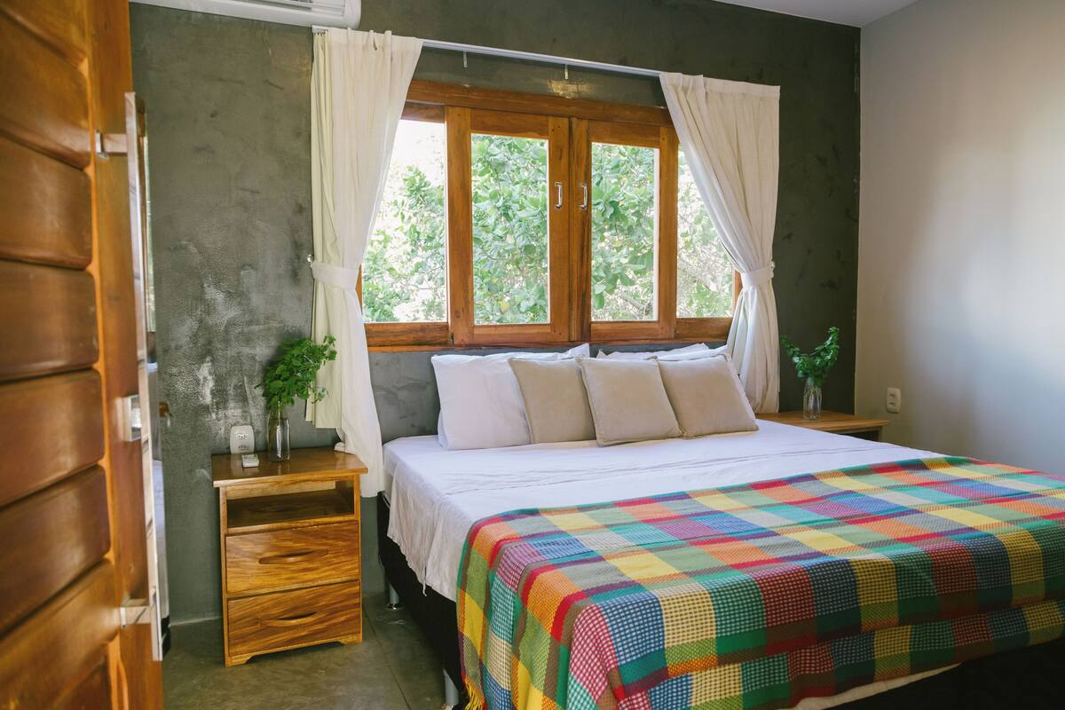 A bedroom with a wooden bed, white bedding, a colorful quilt, a nightstand with a plant, and a window with curtains.