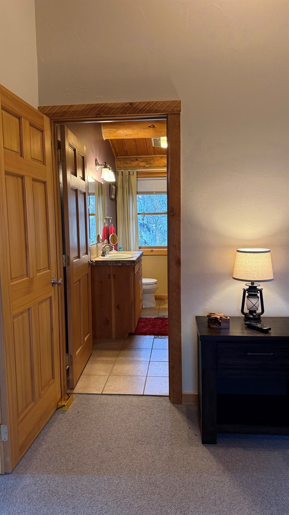 Ein Flur, der zu einem Badezimmer mit Holztür, Waschbecken und Toilette führt. Rechts steht eine Lampe auf einem Tisch.