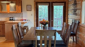 Un coin repas rustique en bois, avec une grande table et des chaises, une cuisine en arrière-plan et une vue sur l’extérieur par une porte.