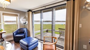 A room with a large sliding glass door leading to a deck with a view of a lake and grassy area. There are two chairs, one blue and one brown, and a small table with a lamp.