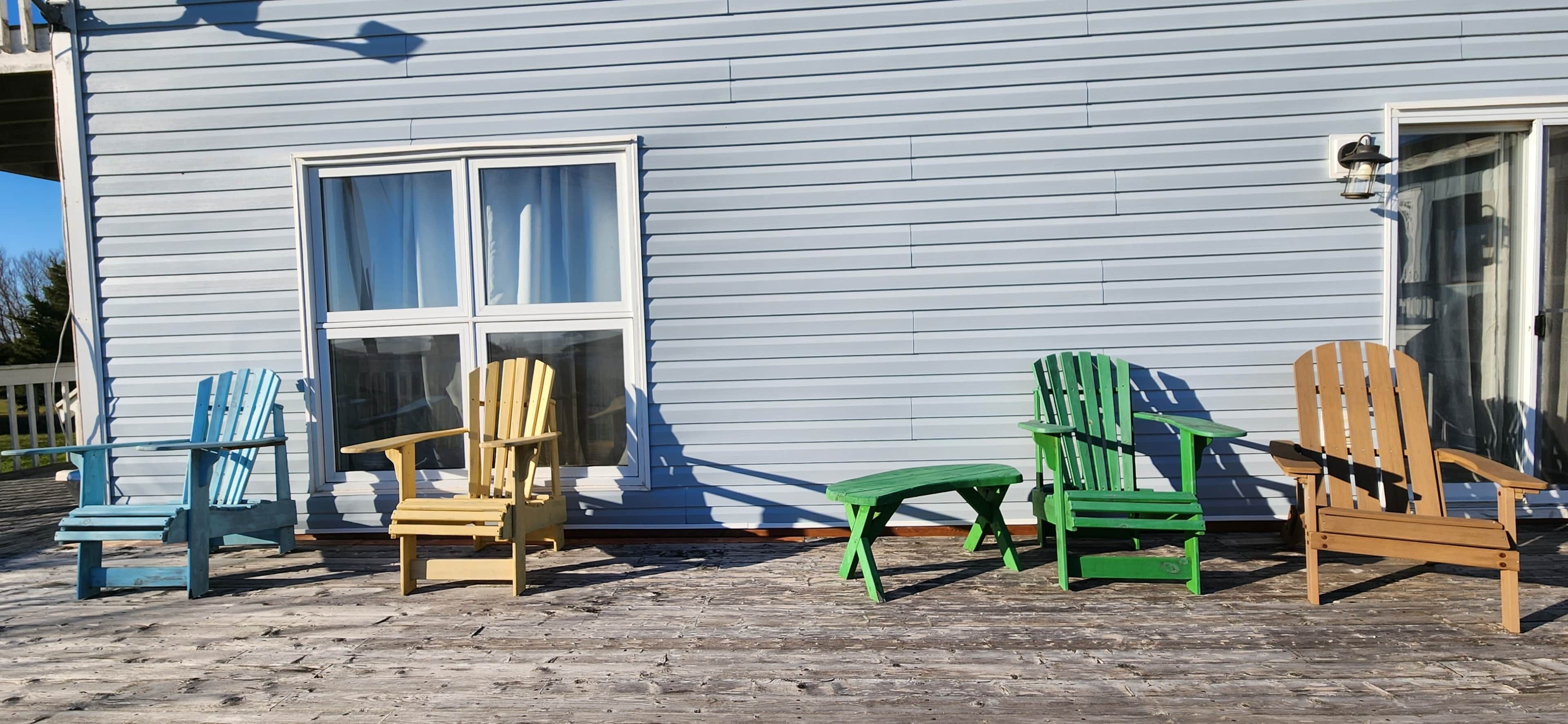 Eine Holzterrasse mit vier Adirondack-Stühlen und einem kleinen Tisch vor einem hellblauen Haus mit einem großen Fenster.