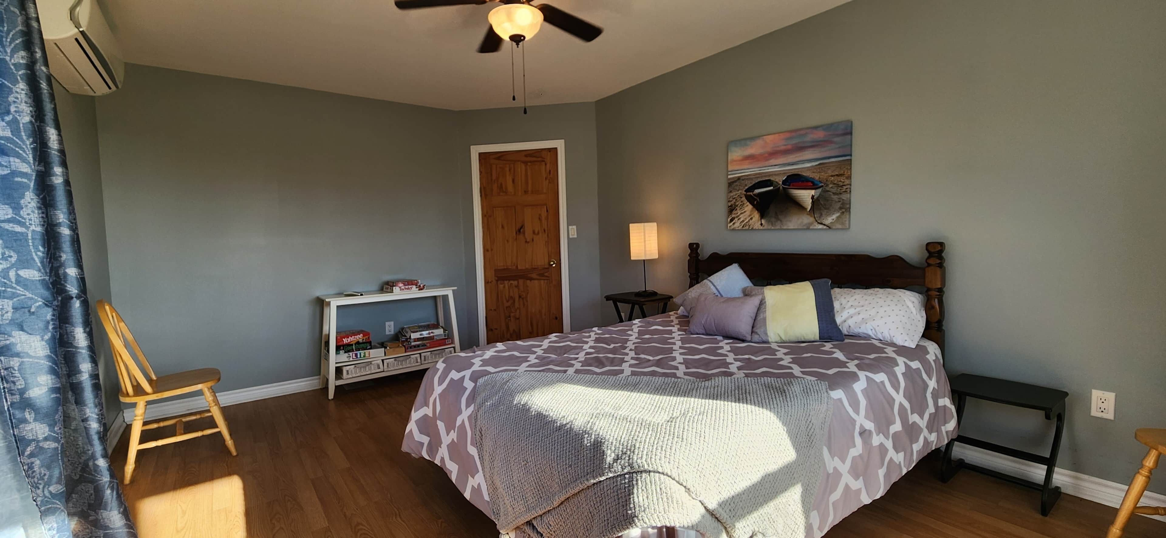 Une chambre à coucher avec un lit, une chaise en bois, une table de chevet, une lampe et un tableau accroché au mur.