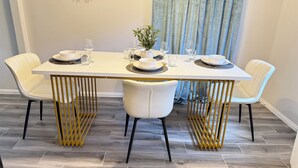 Una mesa de comedor con sillas blancas y un marco dorado, dispuesta con platos, vasos y una planta pequeña.