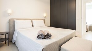 Een net opgemaakt bed met opgerolde handdoeken, een klein nachtkastje en een donkere garderobe.