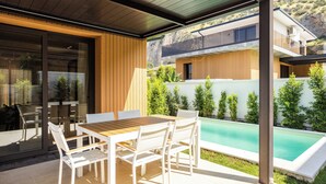 An outdoor patio with a wooden table and white chairs, a swimming pool, and a modern house in the background. - Luxury Villa Artemis (Palermo)
